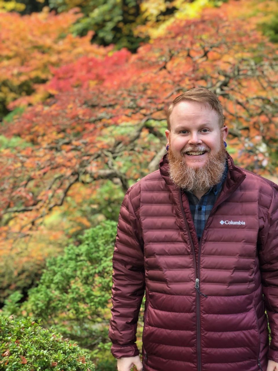 Bradley standing, wearing a marooon columbia jacket in front of colorful autumn foliage at the Portland Japanese Gardens